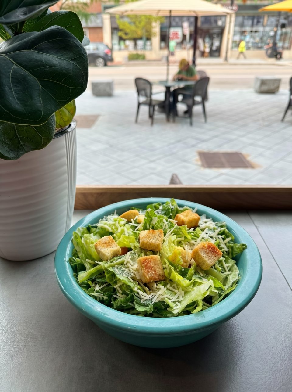 Caesar salad in a turquoise bowl with croutons and cheese, overlooking a street patio.