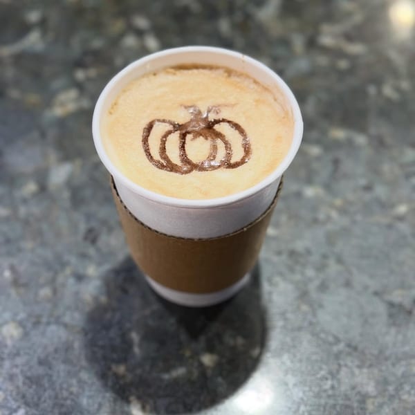 A pumpkin spice latte in a white cup with a brown sleeve, featuring pumpkin latte art.