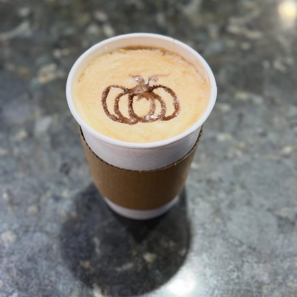 A pumpkin spice latte in a white cup with a brown sleeve, featuring pumpkin latte art.