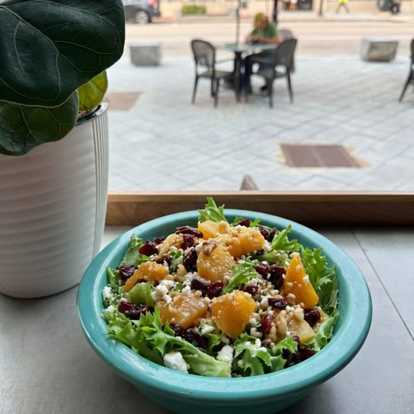 A vibrant salad with lettuce, oranges, cranberries, nuts, and feta cheese in a teal bowl by a window.