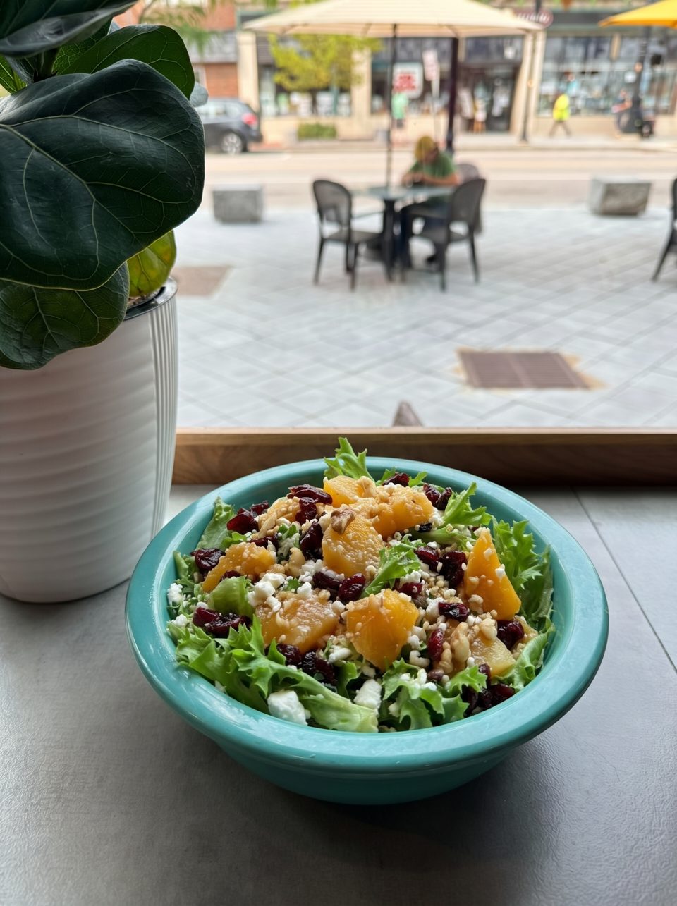 A vibrant salad with lettuce, oranges, cranberries, nuts, and feta cheese in a teal bowl by a window.