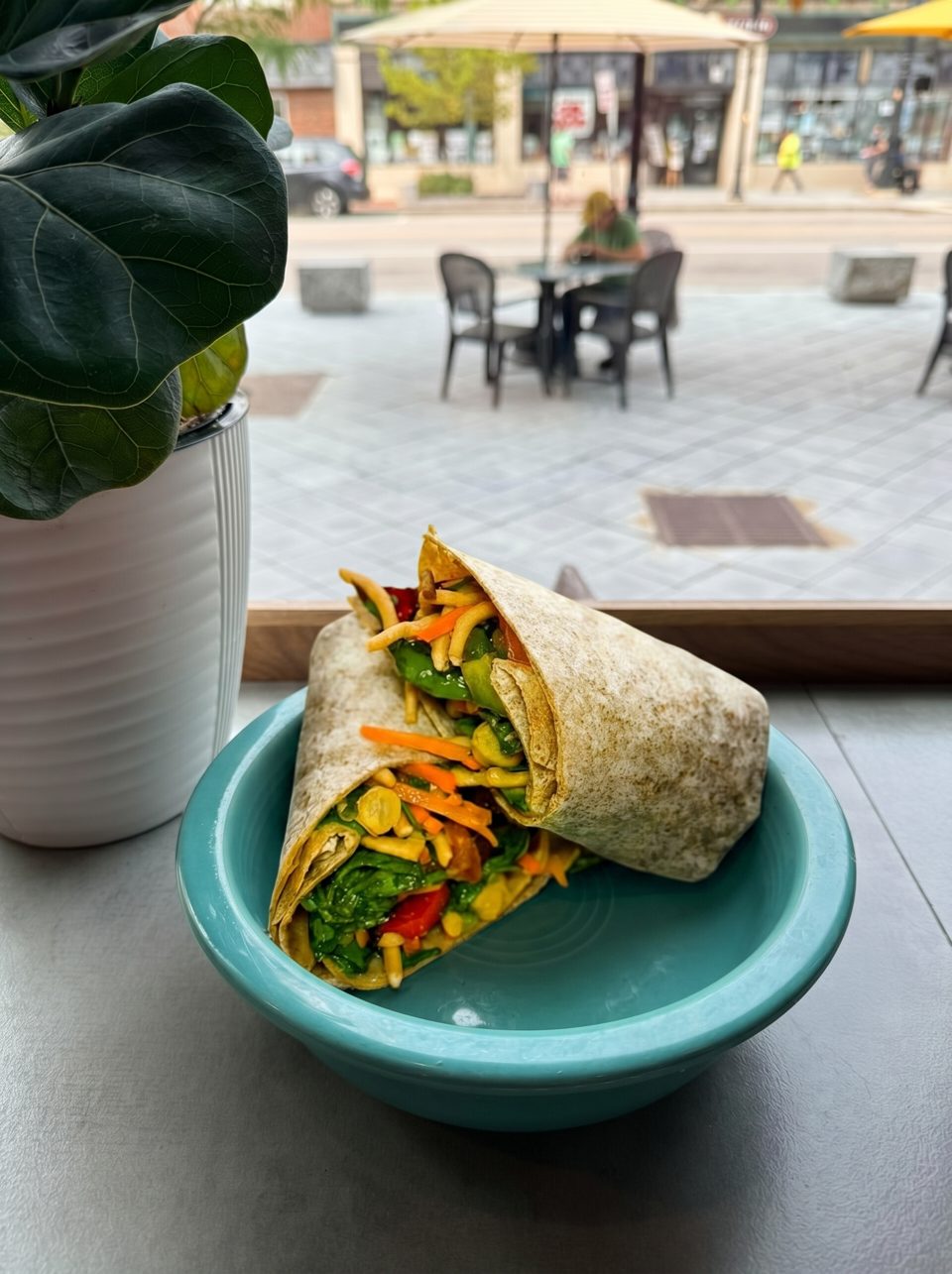 Two colorful wraps filled with vegetables in a teal bowl, with an outdoor street scene in the background.