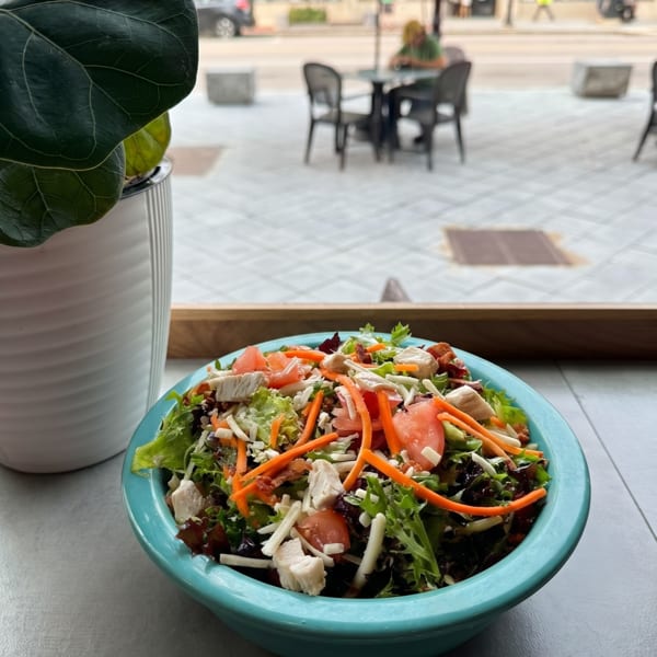 A vibrant salad in a teal bowl with chicken, tomatoes, carrots, and cheese, overlooking a street scene.