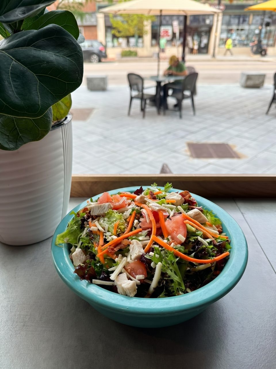 A vibrant salad in a teal bowl with chicken, tomatoes, carrots, and cheese, overlooking a street scene.
